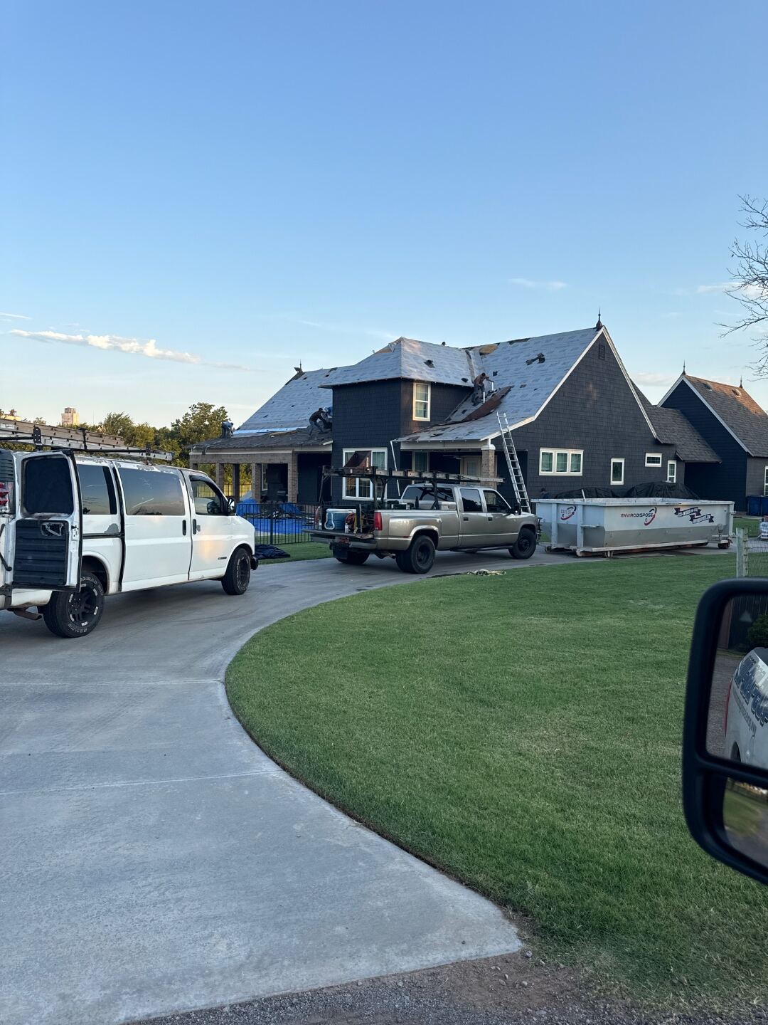 Genesis Roofing crew mid re-roof on a dark-shingle Oklahoma home, with work trucks and dumpster staged in the driveway and a wide open sky behind the house.